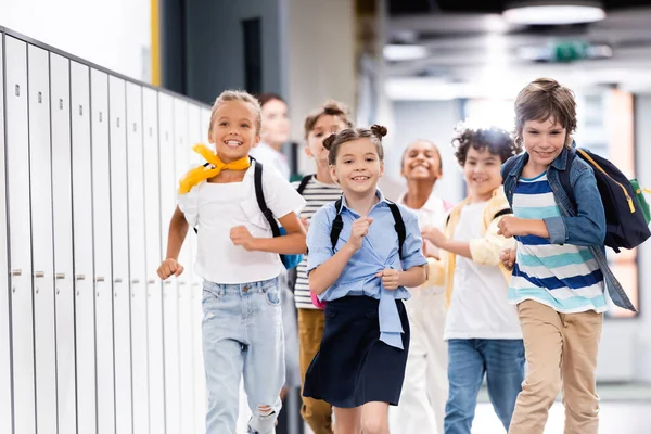 Focus selettivo di compagni di classe multiculturali eccitati con zaini che corrono lungo il corridoio scolastico — Foto stock