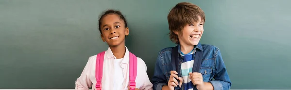 Horizontal concept of african american schoolgirl looking at camera while standing at chalkboard near excited classmate — Stock Photo