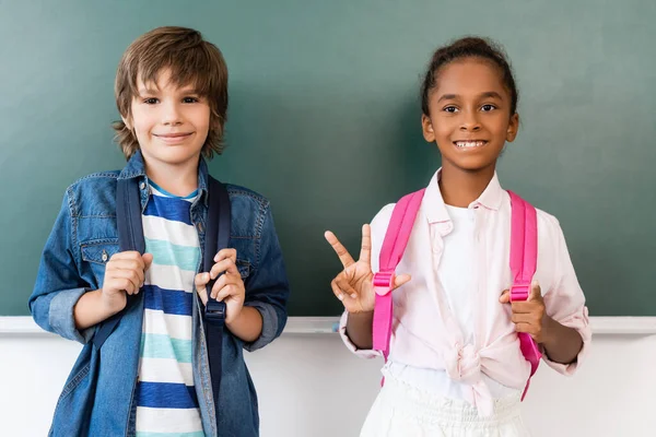 African american schoolgirl showing victory gesture while standing at chalkboard with classmate — Stock Photo