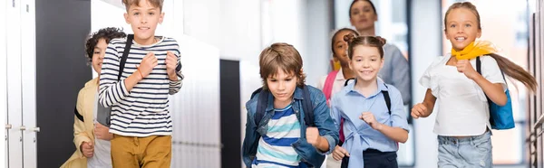 Plano panorámico de compañeros de clase multiétnicos emocionados corriendo por el pasillo de la escuela con el profesor en el fondo — Stock Photo