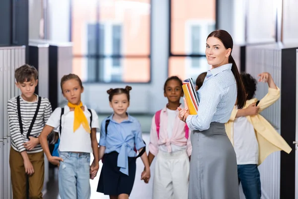 Messa a fuoco selettiva dell'insegnante guardando la telecamera mentre è in piedi con gli alunni multietnici nel corridoio scolastico — Foto stock