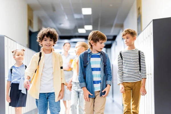 Multicultural schoolchildren walking along school corridor with teacher on background — Stock Photo