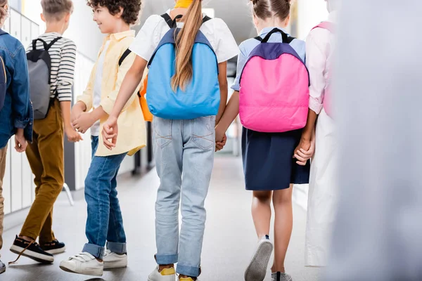 Vista posteriore di compagni di classe multietnici che si tengono per mano mentre camminano lungo il corridoio scolastico, messa a fuoco selettiva — Foto stock