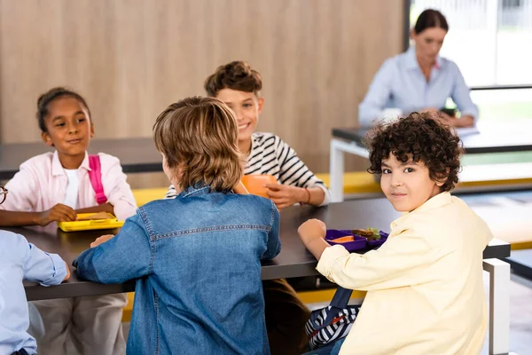 Selective focus of arabian schoolboy looking at camera while sitting in dining room near multicultural classmates and teacher — Stock Photo