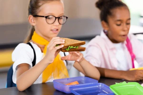 Enfoque selectivo de colegiala en gafas celebración de sándwich cerca de compañero de clase afroamericano en el comedor de la escuela - foto de stock