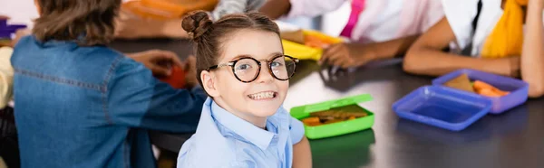 Селективный фокус возбужденной школьницы в очках, смотрящей в камеру рядом с одноклассниками и коробками для обедов, заголовок сайта — стоковое фото