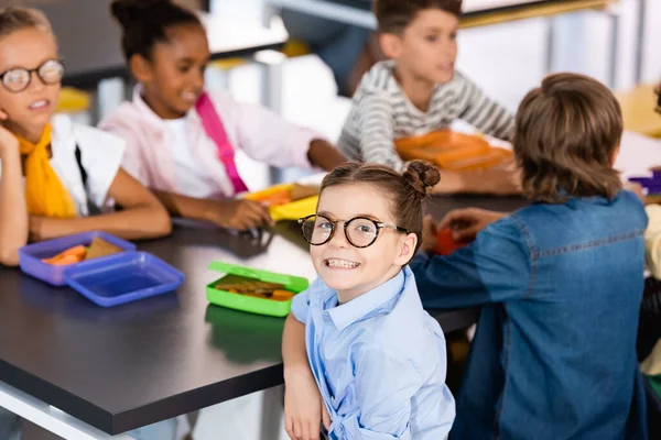 Enfoque selectivo de colegiala emocionada en gafas mirando a la cámara en la cantina de la escuela cerca de compañeros de clase multiétnicos - foto de stock