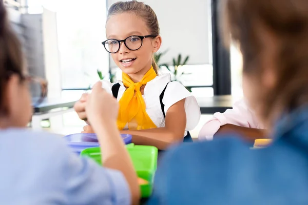 Selective focus of schoolgirl in eyeglasses sitting with classmates in school eatery — Stock Photo