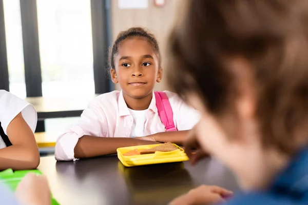 Selective focus of african american schoolgirl sitting in school eatery near lunch box — Stock Photo