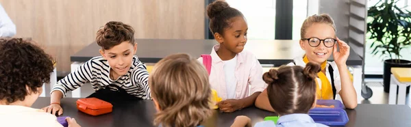 Schülerin berührt Brille, während sie mit multiethnischen Klassenkameraden in der Schulkantine sitzt, horizontales Bild — Stockfoto