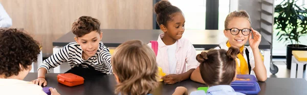 Colegiala tocar gafas mientras está sentado con compañeros de clase multiétnicos en la cantina de la escuela, imagen horizontal - foto de stock