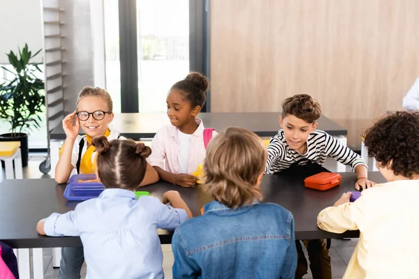 Schoolgirl touching eyeglasses while sitting with multicultural classmates in school eatery — Stock Photo