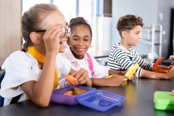 Foyer sélectif de l'écolière touchant les lunettes et tenant la carotte fraîche dans la cantine de l'école près des amis multiculturels — Photo de stock