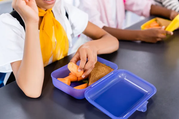 Обрезанный вид школьницы, берущей свежую морковку из коробки для ланча рядом с африканским одноклассником — стоковое фото