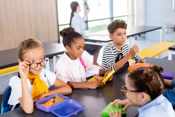 Colegialas hablando mientras están sentadas en el comedor de la escuela con compañeros de clase multiculturales y profesor de fondo - foto de stock