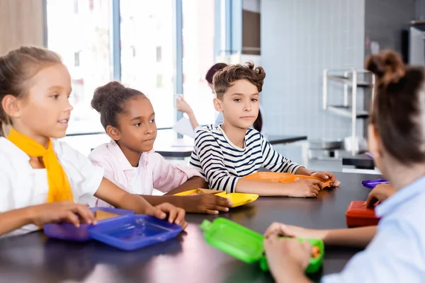 Focus selettivo degli alunni multiculturali seduti nella mensa scolastica vicino ai cestini del pranzo — Foto stock