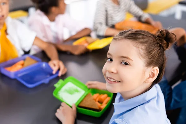 Foyer sélectif de l'écolière regardant la caméra près de la boîte à lunch avec sandwich et carottes fraîches — Photo de stock
