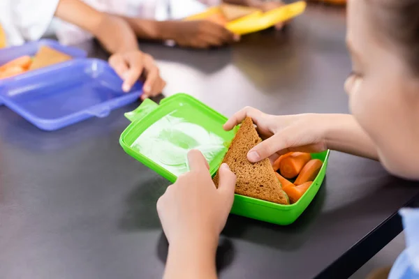 Selective focus of schoolgirl taking sandwich from lunch box with fresh carrots near classmates — Stock Photo