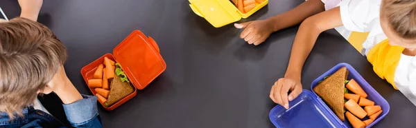 Vue aérienne des élèves multiculturels près des boîtes à lunch avec sandwichs et carottes fraîches, image horizontale — Photo de stock