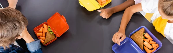 Vista aérea de alumnos multiculturales cerca de loncheras con sándwiches y zanahorias frescas, imagen horizontal - foto de stock