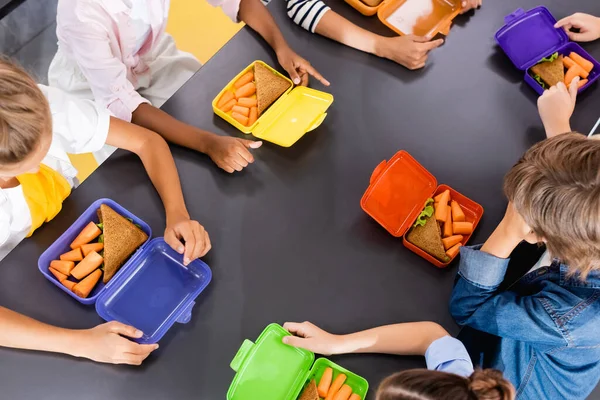 Overhead view of multicultural classmates sitting in school eatery near lunch boxes with fresh carrots and sandwiches — Stock Photo