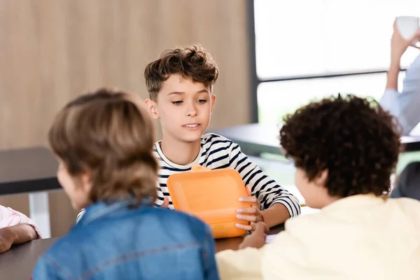Селективное внимание школьника, открывающего коробку с обедом, сидящего в школьной столовой рядом с одноклассниками — стоковое фото
