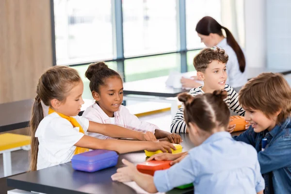 Focus selettivo dei compagni di classe multietnici seduti nella mensa scolastica vicino ai cestini del pranzo — Foto stock