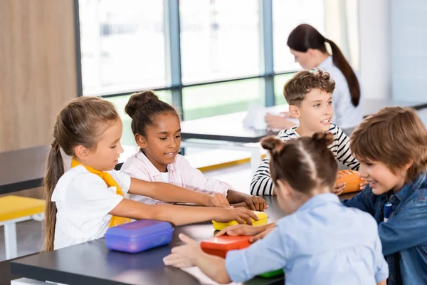 Enfoque selectivo de compañeros de clase multiétnicos sentados en la cantina de la escuela cerca de loncheras - foto de stock