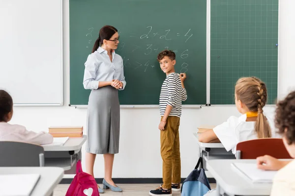 Colegial mirando a compañeros de clase multiculturales mientras resuelve ecuaciones en pizarra durante la lección de matemáticas - foto de stock