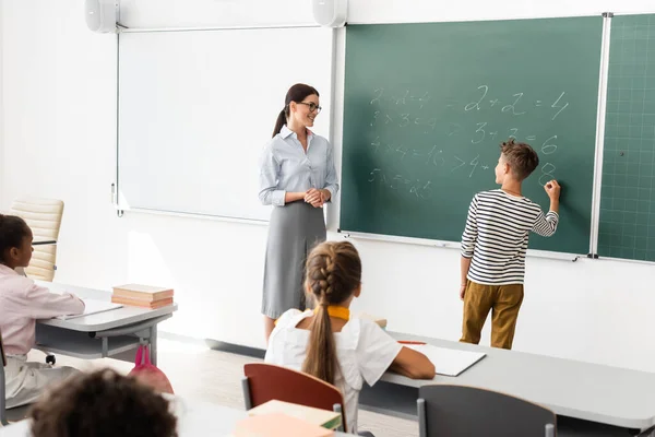Vue arrière des équations de résolution des écoliers sur tableau près de l'enseignant et des élèves multiethniques — Photo de stock