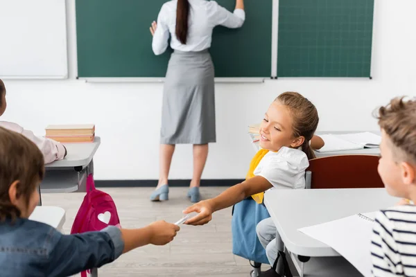 Selective focus of pupil passing note to schoolgirl while teacher writing on chalkboard during lesson — Stock Photo