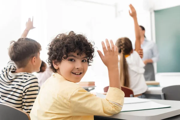 Selective focus of arabian schoolboy with hand in air looking at camera near multicultural classmates — Stock Photo