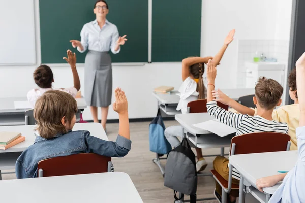 Vista traseira de alunos multiculturais com as mãos no ar, e professor de pé com os braços abertos perto de quadro-negro em sala de aula — Fotografia de Stock