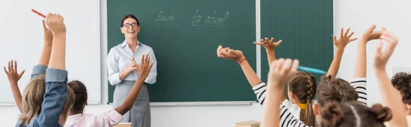 Back view of multicultural pupils with hands in air, and teacher standing at chalkboard with back to school inscription, horizontal image — Stock Photo