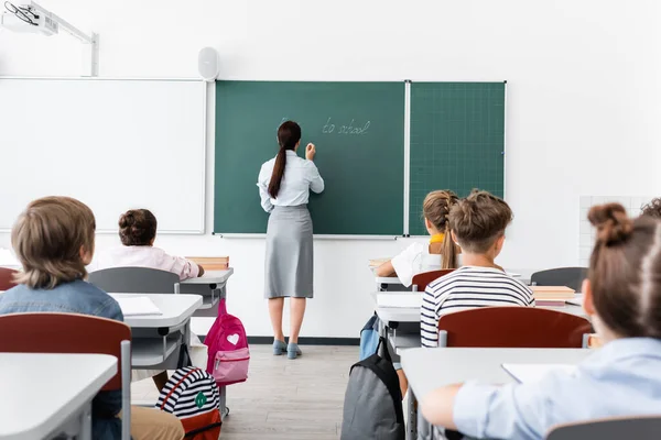 Back view of teacher in formal wear writing on chalkboard near multiethnic pupils in classroom — Stock Photo