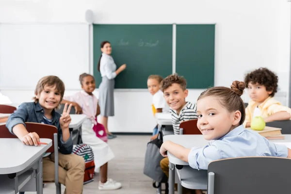 Enfoque selectivo de los alumnos multiculturales mirando a la cámara, y el profesor de pie en la pizarra en el fondo - foto de stock