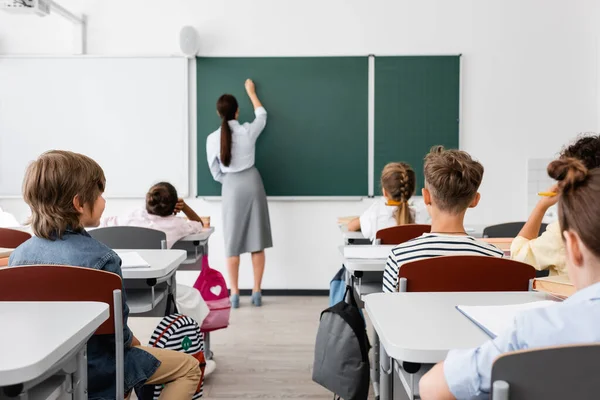 Back view of teacher writing on chalkboard near multicultural pupils in classroom — Stock Photo