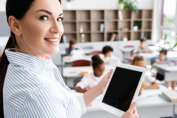 Selective focus of teacher holding digital tablet with blank screen and looking at camera near multicultural pupils in classroom — Stock Photo