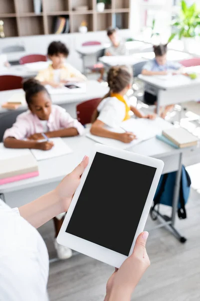 Vista parcial del profesor sosteniendo tableta digital con pantalla en blanco cerca de alumnos multiculturales escribiendo durante la lección - foto de stock