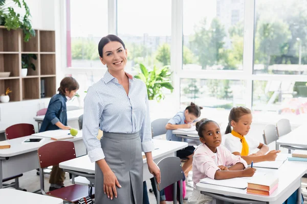 Lehrer blickt in Kamera, während er im Klassenzimmer in der Nähe multikultureller Schüler steht — Stockfoto