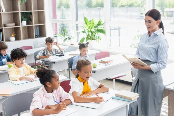 Selective focus of teacher with book talking to african american girl near multicultural pupils during lesson — Stock Photo