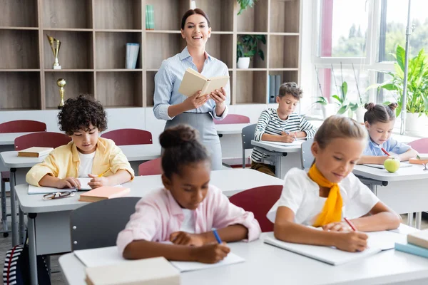 Teacher holding book and looking at camera while multicultural pupils writing dictation in classroom — Stock Photo
