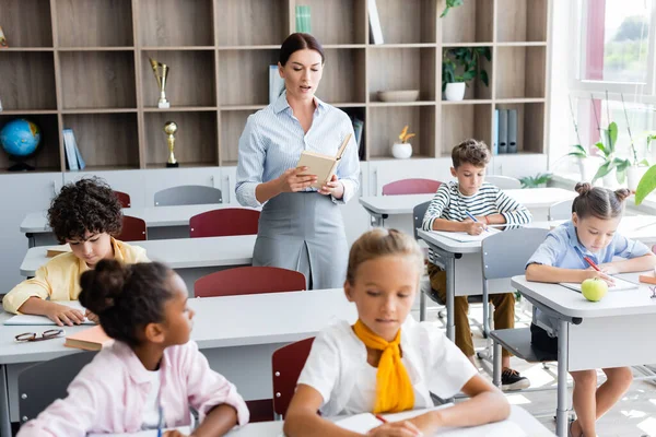 Enfoque selectivo del libro de lectura del profesor mientras los alumnos multiculturales escriben dictado en el aula — Stock Photo