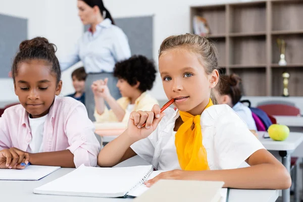 Selektiver Fokus einer Schülerin, die in der Nähe eines afrikanisch-amerikanischen Freundes am Schreibtisch in der Schule in die Kamera blickt — Stockfoto