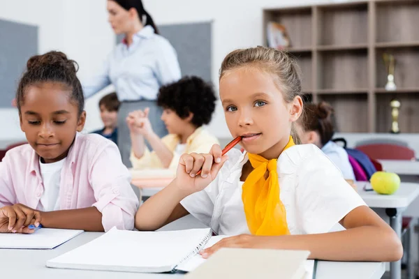 Focus selettivo della studentessa che guarda la fotocamera vicino all'amico afro-americano alla scrivania a scuola — Foto stock