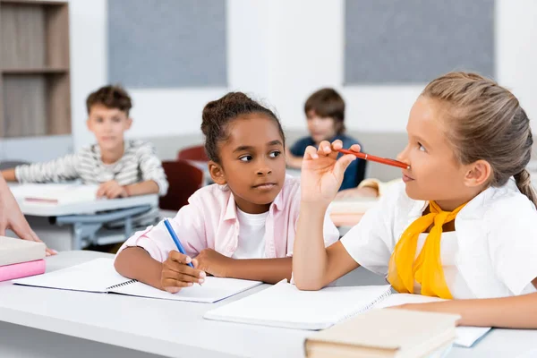 Selective focus of multiethnic schoolgirls writing on notebooks during lesson in classroom — Stock Photo