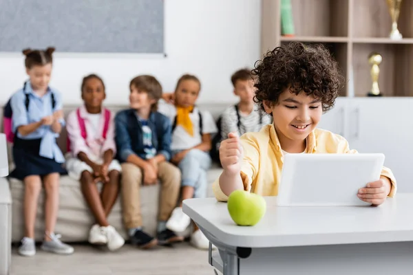 Selective focus of muslim schoolboy using digital tablet near apple on desk in classroom — Stock Photo