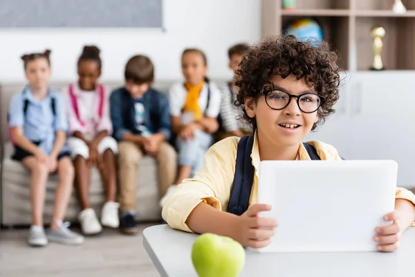 Selective focus of muslim schoolboy using digital tablet near apple on desk and multiethnic classmates at background — Stock Photo