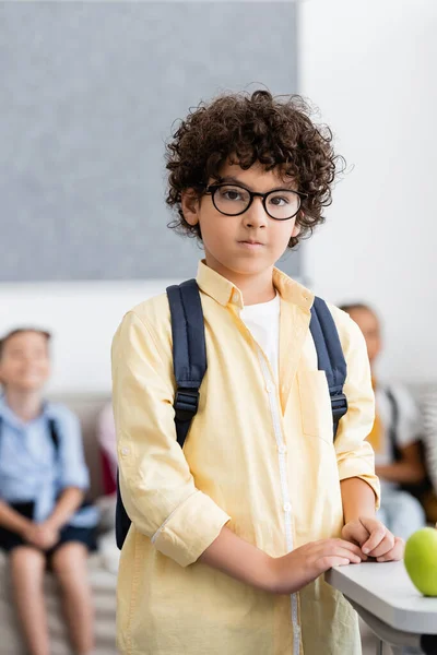 Selective focus of muslim schoolboy with backpack standing near apple on desk in classroom — Stock Photo