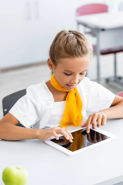 Selective focus of schoolgirl using digital tablet near apple on desk in classroom — Stock Photo